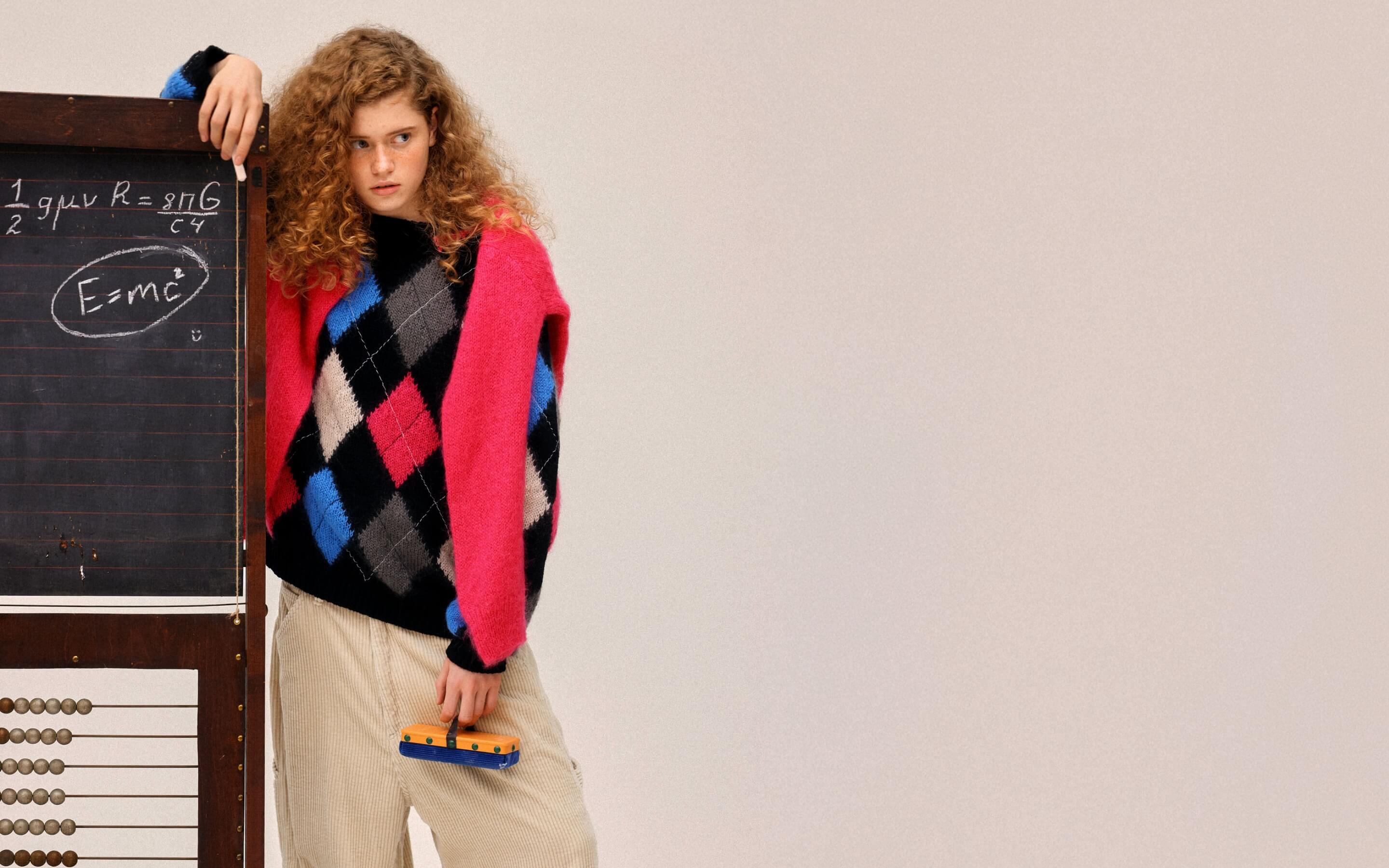A model wearing an argyle sweater and pink cardigan leaning against a chalkboard with equations.