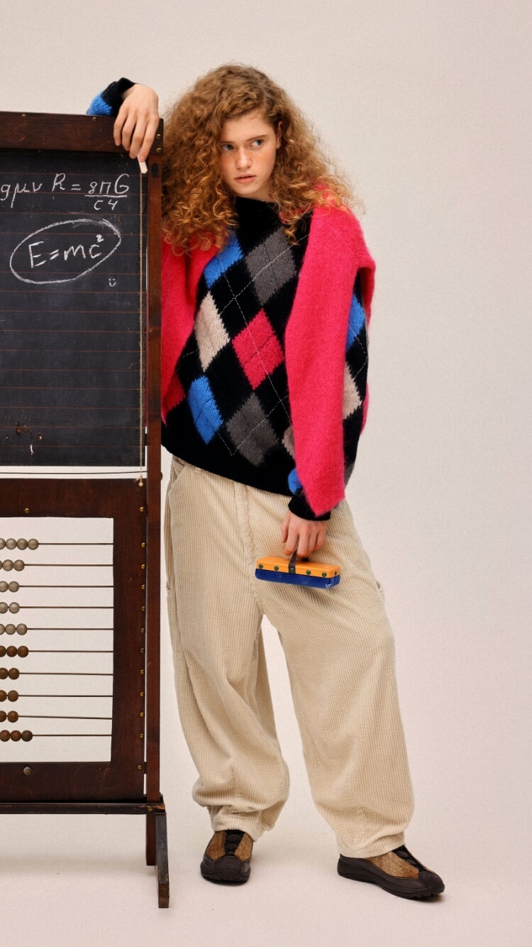 A model wearing an argyle sweater and pink cardigan leaning against a chalkboard with equations.